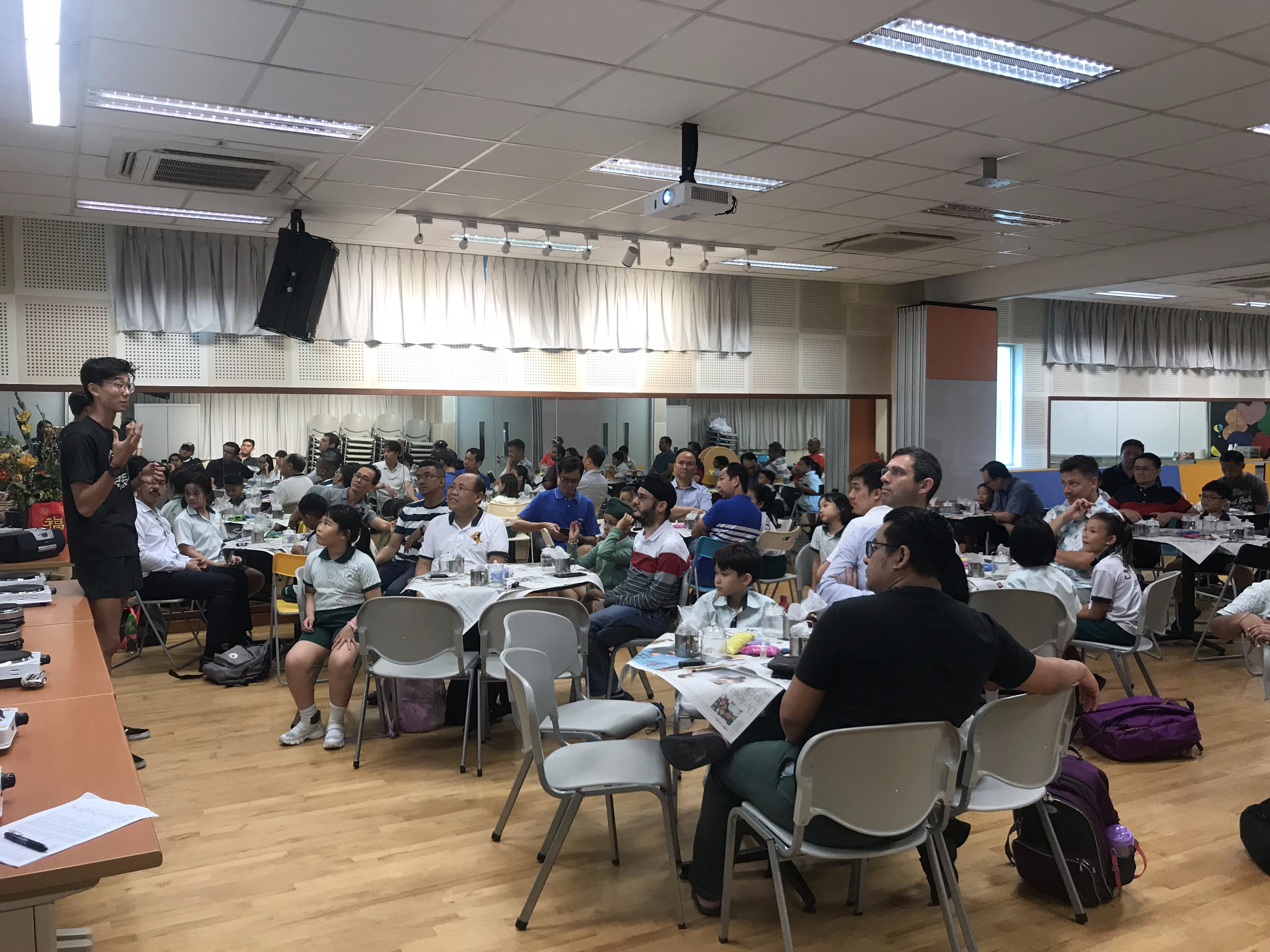 A person stands speaking to a large group of adults and children seated at tables in a school hall.